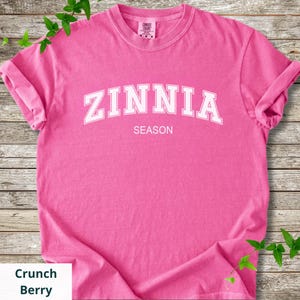 May include: A vibrant pink t-shirt displaying the text "ZINNIA SEASON" in white, bold lettering. The shirt has rolled-up sleeves and is a solid colour. The label "Crunch Berry" is in a white box.