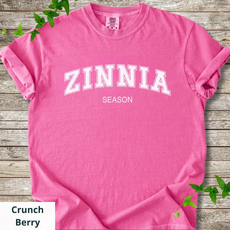 May include: A vibrant pink t-shirt displaying the text "ZINNIA SEASON" in white, bold lettering. The shirt has rolled-up sleeves and is a solid colour. The label "Crunch Berry" is in a white box.