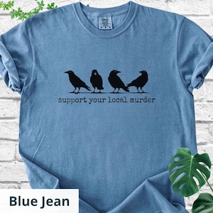 May include: A blue jean colored short-sleeve t-shirt featuring a black graphic of four crows standing in a row. Below the birds, the text reads "support your local murder" in a typewriter-style font. The shirt has a crew neckline and a tag.