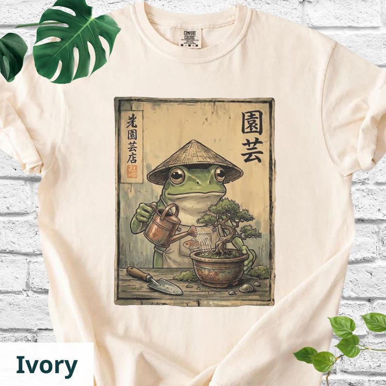 May include: Ivory-colored t-shirt featuring a frog wearing a conical hat, watering a bonsai tree. The frog is depicted in a vintage style, with Japanese text above and to the left of the frog. A small shovel is at the bottom.