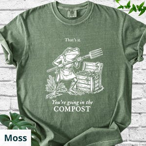 May include: Moss green t-shirt with a humorous graphic. The design features a frog wearing an apron and holding a pitchfork, with the text "That's it." and "You're going in the COMPOST."