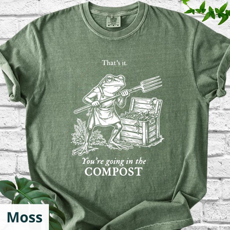 May include: Moss green t-shirt with a humorous graphic. The design features a frog wearing an apron and holding a pitchfork, with the text "That's it." and "You're going in the COMPOST."