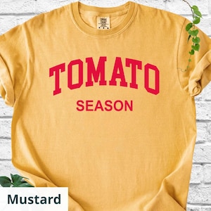 K&ouml;nnte beinhalten: Senffarbenes T-Shirt mit dem roten Aufdruck "TOMATO SEASON". Das T-Shirt hat einen Rundhalsausschnitt und kurze &Auml;rmel. Der Stoff wirkt weich und bequem.