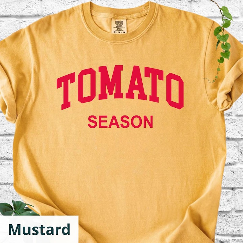 K&ouml;nnte beinhalten: Senffarbenes T-Shirt mit dem roten Aufdruck "TOMATO SEASON". Das T-Shirt hat einen Rundhalsausschnitt und kurze &Auml;rmel. Der Stoff wirkt weich und bequem.