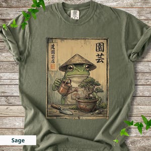 May include: Sage green t-shirt featuring a frog wearing a conical hat, watering a bonsai tree. The frog is depicted in a Japanese art style. The shirt has Japanese text and the word "Sage" at the bottom.