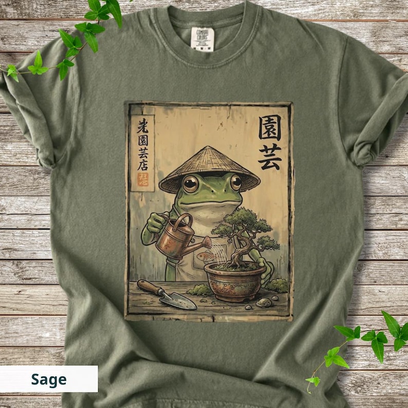 May include: Sage green t-shirt featuring a frog wearing a conical hat, watering a bonsai tree. The frog is depicted in a Japanese art style. The shirt has Japanese text and the word "Sage" at the bottom.