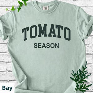 K&ouml;nnte beinhalten: Hellgr&uuml;nes T-Shirt mit dem Aufdruck "TOMATO SEASON" in dunkelgr&uuml;nen Blockbuchstaben. Das Shirt besteht aus weichem Material und hat hochgekrempelte &Auml;rmel. Das Shirt wird vor einer wei&szlig;en Backsteinmauer pr&auml;sentiert.
