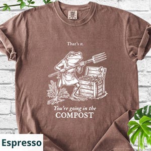 May include: Espresso-colored t-shirt featuring a humorous graphic of a frog holding a pitchfork, standing near a compost bin. The text reads "That's it. You're going in the COMPOST."