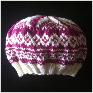 Könnte beinhalten: Eine gestrickte Mütze mit Fair-Isle-Muster in Magenta und Weiß. Die Mütze hat ein sich wiederholendes geometrisches Design. Der Rand ist einfarbig weiß. Die Mütze ist aus einem weichen, warmen Material gefertigt, ideal für kaltes Wetter.