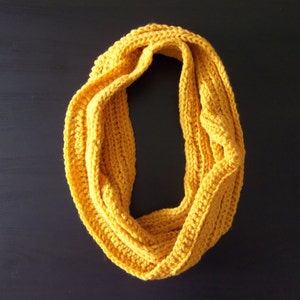Könnte beinhalten: Senfgelber Loop-Schal, handgehäkelt mit Rippenstruktur. Der Schal ist aus weichem Garn gefertigt und vor einem dunklen Hintergrund dargestellt. Dieses gemütliche Accessoire ist perfekt, um Wärme und Stil zu verleihen.