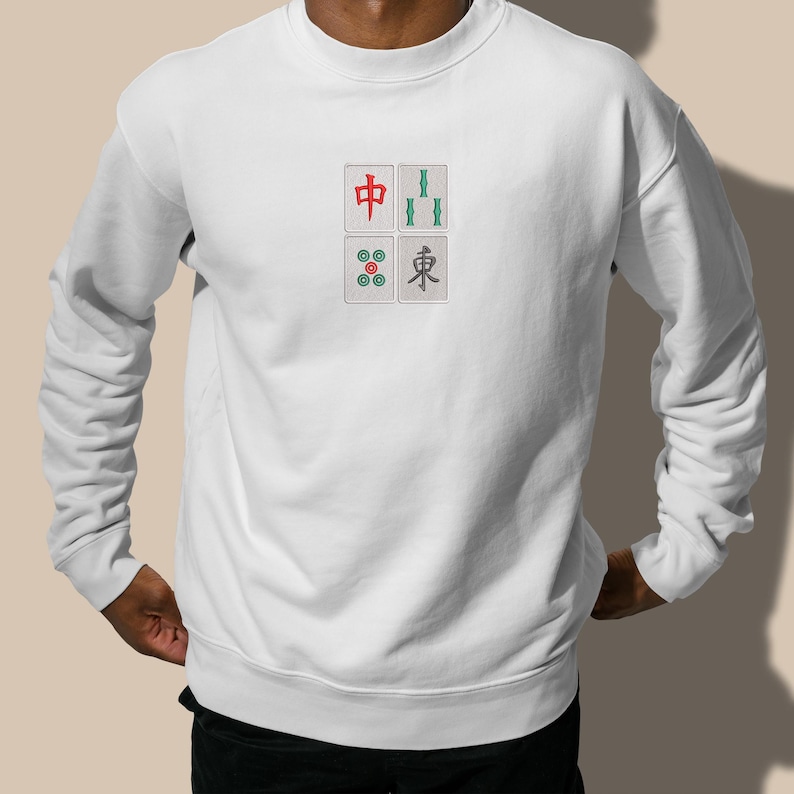 May include: White sweatshirt with a four-tile Mahjong design embroidered on the front. The design includes red, green, and blue details. The sweatshirt is a casual, long-sleeved garment.