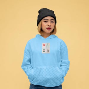 May include: Light blue hoodie with a front pocket and a small embroidered design of four Mahjong tiles. The hoodie has a drawstring hood and long sleeves. The person in the image is wearing a black beanie.