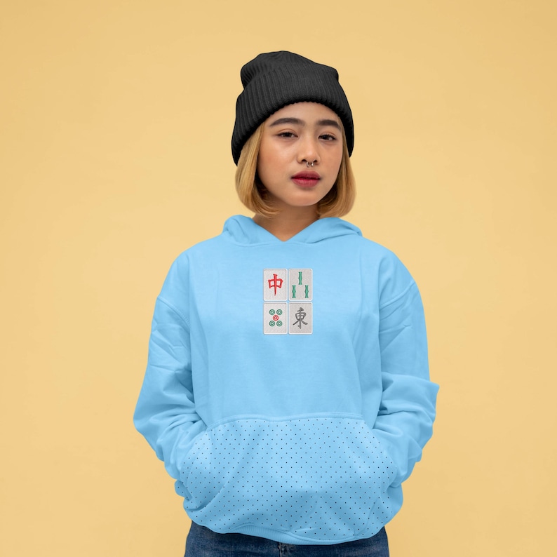 May include: Light blue hoodie with a front pocket and a small embroidered design of four Mahjong tiles. The hoodie has a drawstring hood and long sleeves. The person in the image is wearing a black beanie.