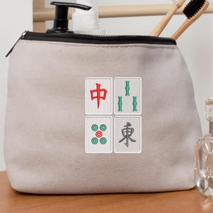 May include: A beige canvas cosmetic bag with a black zipper and four embroidered Mahjong tile designs. The tiles feature red, green, and teal symbols. The bag is on a wooden surface.