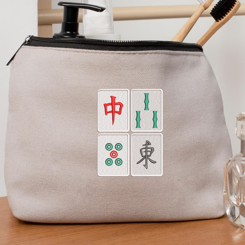May include: A beige canvas cosmetic bag with a black zipper and four embroidered Mahjong tile designs. The tiles feature red, green, and teal symbols. The bag is on a wooden surface.