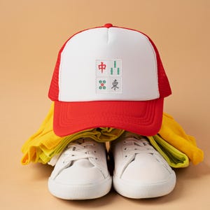 May include: A red and white trucker hat with a white front panel featuring embroidered Mahjong tiles. The hat is paired with white sneakers and yellow clothing, set against a neutral background. The hat's design includes Chinese characters.
