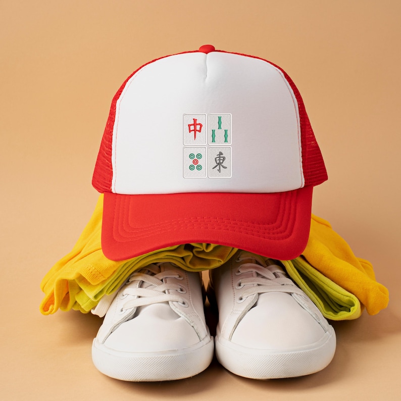 May include: A red and white trucker hat with a white front panel featuring embroidered Mahjong tiles. The hat is paired with white sneakers and yellow clothing, set against a neutral background. The hat's design includes Chinese characters.