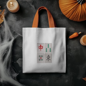 May include: White tote bag with orange handles, featuring four embroidered Mahjong tiles. The tiles display Chinese characters and symbols in red, green, and black. The bag is set against a dark background with Halloween decorations.