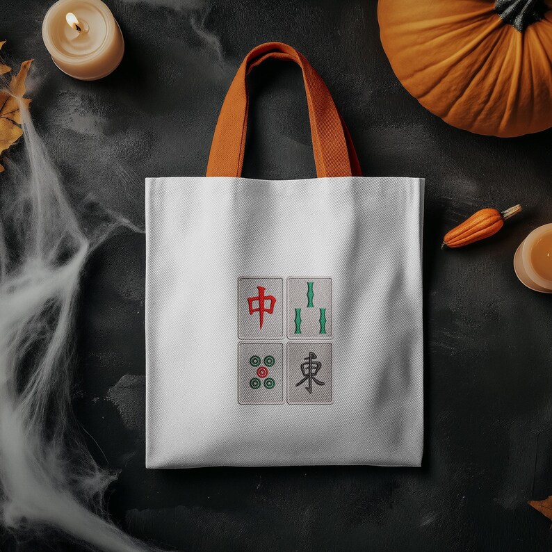 May include: White tote bag with orange handles, featuring four embroidered Mahjong tiles. The tiles display Chinese characters and symbols in red, green, and black. The bag is set against a dark background with Halloween decorations.
