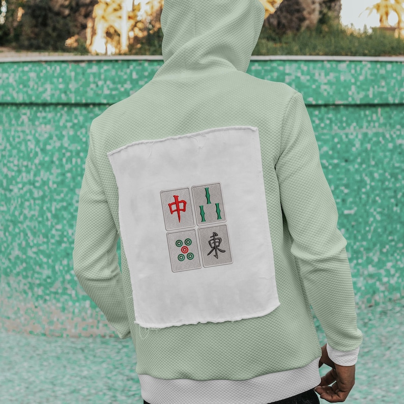 May include: A light green hoodie with a white patch on the back featuring a Mahjong tile design. The design includes red, green, and gray tiles with Chinese characters. The hoodie has a hood and a textured fabric.
