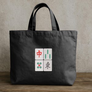 May include: Black tote bag with four embroidered Mahjong tile designs. The tiles feature red, green, and blue symbols on a white background. The bag has black handles and is made of a sturdy fabric.