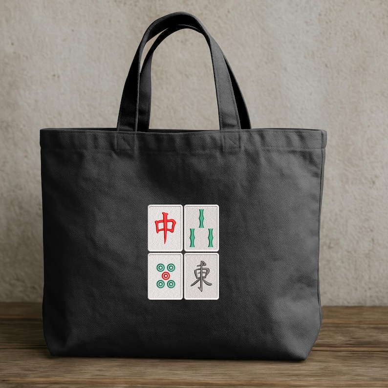 May include: Black tote bag with four embroidered Mahjong tile designs. The tiles feature red, green, and blue symbols on a white background. The bag has black handles and is made of a sturdy fabric.