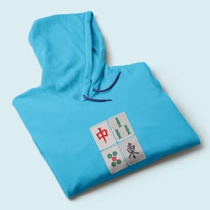 May include: A light blue hoodie with a drawstring hood. The hoodie features four embroidered Mahjong tile designs in red, green, and black. The hoodie is made of a soft material.