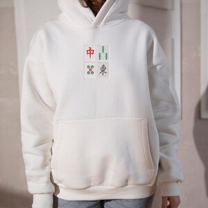 May include: Cream-colored hoodie with a front pocket and a hood. The front features four small squares with Chinese characters and symbols in red, green, and gray. A casual, comfortable garment.