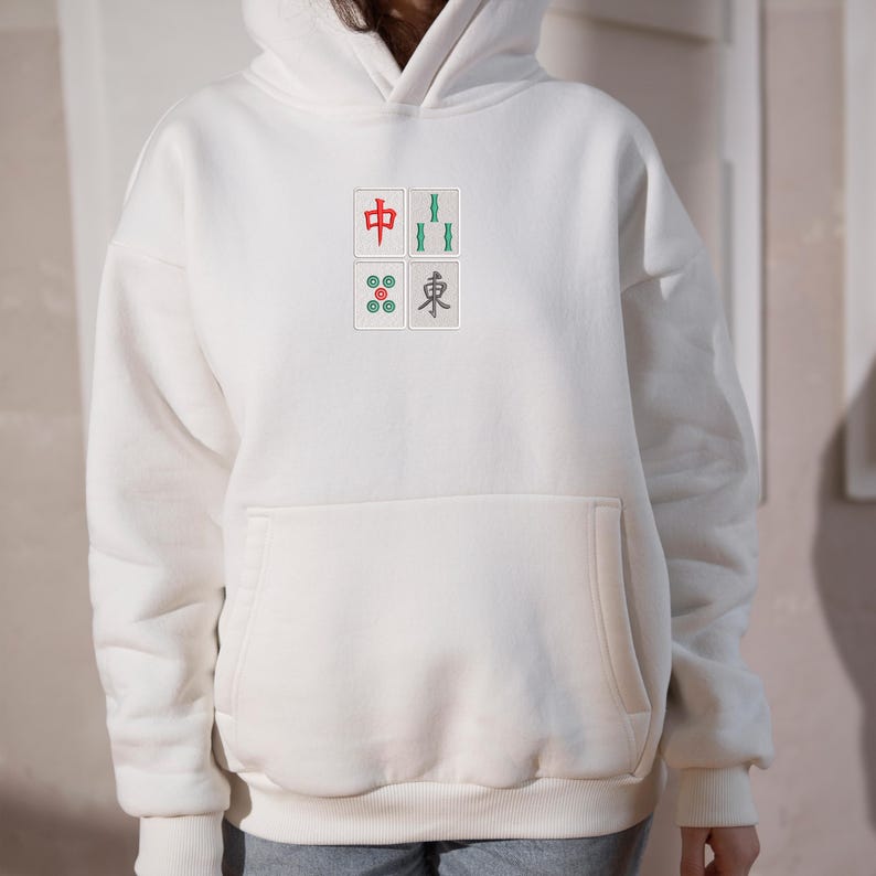 May include: Cream-colored hoodie with a front pocket and a hood. The front features four small squares with Chinese characters and symbols in red, green, and gray. A casual, comfortable garment.