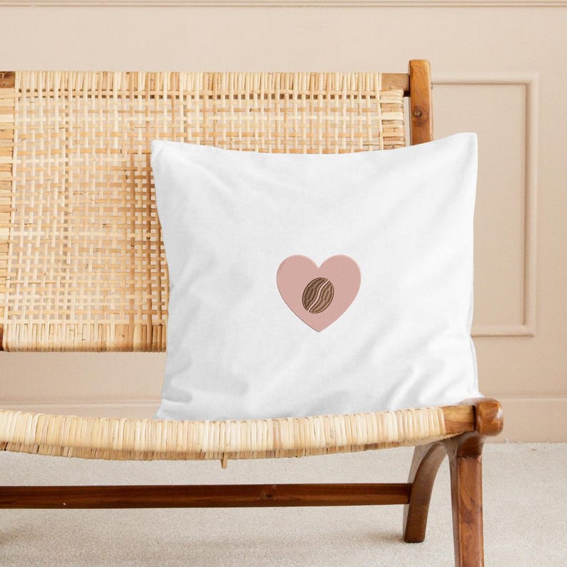 Puede incluir: Coj&iacute;n cuadrado blanco con un dise&ntilde;o de coraz&oacute;n rosa y una ilustraci&oacute;n de grano de caf&eacute;. El coj&iacute;n est&aacute; colocado sobre una silla tejida con un marco de madera marr&oacute;n claro. Art&iacute;culo decorativo.