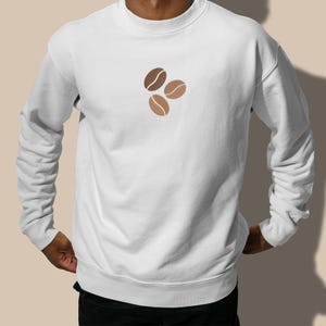 Puede incluir: Sudadera blanca con cuello redondo y mangas largas. La parte delantera presenta un dise&ntilde;o de tres granos de caf&eacute; en tonos marrones. La sudadera est&aacute; hecha de un material suave.