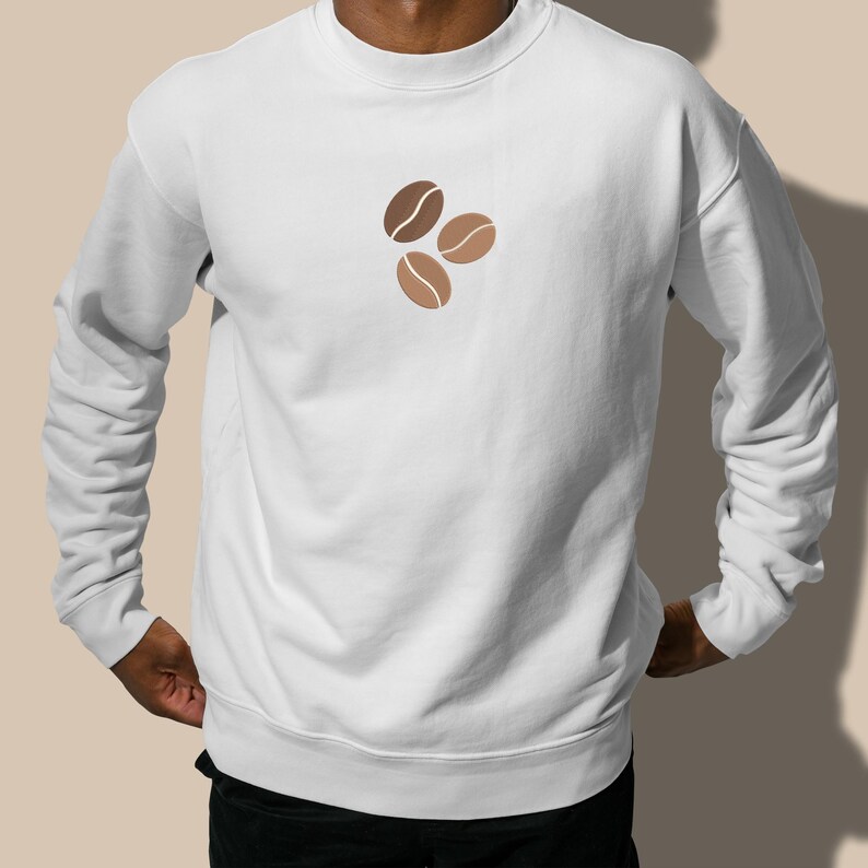 Puede incluir: Sudadera blanca con cuello redondo y mangas largas. La parte delantera presenta un dise&ntilde;o de tres granos de caf&eacute; en tonos marrones. La sudadera est&aacute; hecha de un material suave.