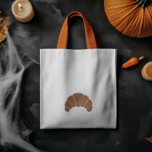 Puede incluir: Bolso tote blanco con un dise&ntilde;o de croissant marr&oacute;n y asas naranjas. El bolso est&aacute; sobre un fondo oscuro con decoraciones de Halloween, incluyendo una calabaza y una vela.