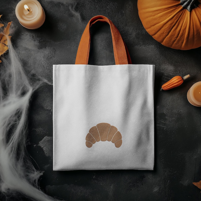 Puede incluir: Bolso tote blanco con un dise&ntilde;o de croissant marr&oacute;n y asas naranjas. El bolso est&aacute; sobre un fondo oscuro con decoraciones de Halloween, incluyendo una calabaza y una vela.