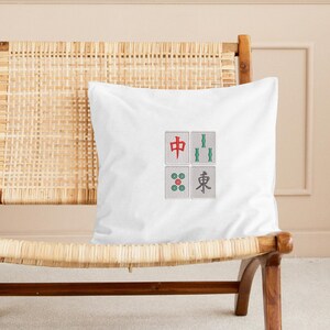 May include: White square pillow featuring four embroidered Mahjong tile designs. The tiles include a red character, green bamboo, a circle design, and a character in gray. The pillow is on a woven chair.