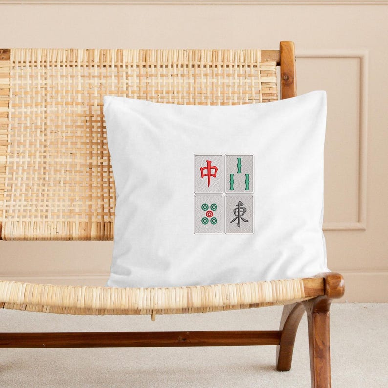May include: White square pillow featuring four embroidered Mahjong tile designs. The tiles include a red character, green bamboo, a circle design, and a character in gray. The pillow is on a woven chair.