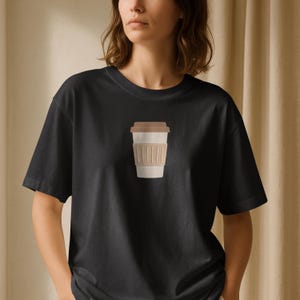 Puede incluir: Camiseta negra con un gr&aacute;fico de taza de caf&eacute;. El gr&aacute;fico muestra una taza beige con tapa marr&oacute;n y una funda beige. La camiseta est&aacute; hecha de un material suave y tiene cuello redondo cl&aacute;sico.