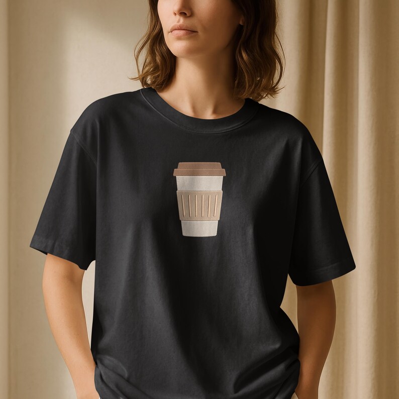 Puede incluir: Camiseta negra con un gr&aacute;fico de taza de caf&eacute;. El gr&aacute;fico muestra una taza beige con tapa marr&oacute;n y una funda beige. La camiseta est&aacute; hecha de un material suave y tiene cuello redondo cl&aacute;sico.