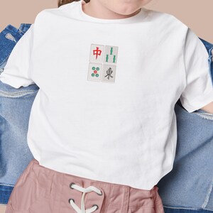 May include: White t-shirt featuring a Mahjong tile design. The design includes four tiles with Chinese characters and symbols in red, green, and blue. The shirt is made of a soft material and is suitable for casual wear.