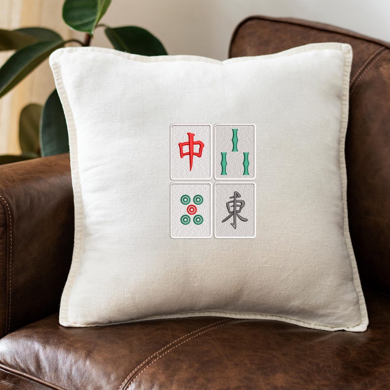 May include: Cream-colored square pillow with a Mahjong tile design. The design features four embroidered tiles with red, green, and gray symbols. The pillow is placed on a brown leather couch.