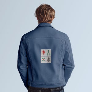 May include: A denim jacket in a medium blue color. The back of the jacket features four embroidered Mahjong tiles in red, green, and white. The jacket has a classic design with a button-up front and pockets.