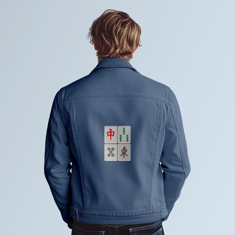 May include: A denim jacket in a medium blue color. The back of the jacket features four embroidered Mahjong tiles in red, green, and white. The jacket has a classic design with a button-up front and pockets.