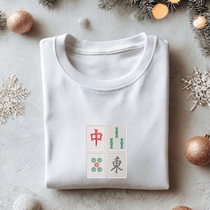 May include: White crew neck t-shirt with four embroidered Mahjong tile designs. The tiles feature red, green, and teal accents. The shirt is folded neatly on a light gray surface, with holiday decorations in the background.