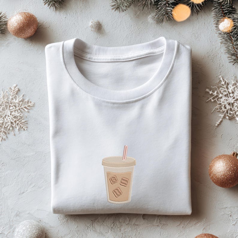 Puede incluir: Una camiseta blanca de cuello redondo con un gr&aacute;fico de caf&eacute; helado. La bebida est&aacute; en un vaso transparente con cubitos de hielo y una pajita rosa. La camiseta est&aacute; doblada sobre una superficie gris claro con adornos navide&ntilde;os.