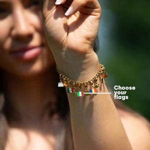 Puede incluir: Pulsera dorada con colgantes de banderas. La pulsera se lleva en la muñeca, con el texto "Choose your flags" visible. Los colgantes incluyen banderas de diferentes países.