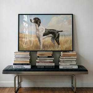 Op de afbeelding: Een ingelijst hondenportret hangt boven een zwarte bank met stapels boeken. De bank heeft een metalen frame en een gecapitonneerde zitting. De boeken bevatten titels als "Modernism" en "Design as Art".