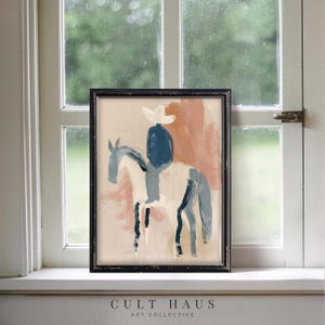 May include: Abstract art print of a cowboy on horseback, rendered in muted tones of blue, white, and peach. The artwork is framed in a black wooden frame. The text "CULT HAUS ART COLLECTIVE" is printed at the bottom of the frame.