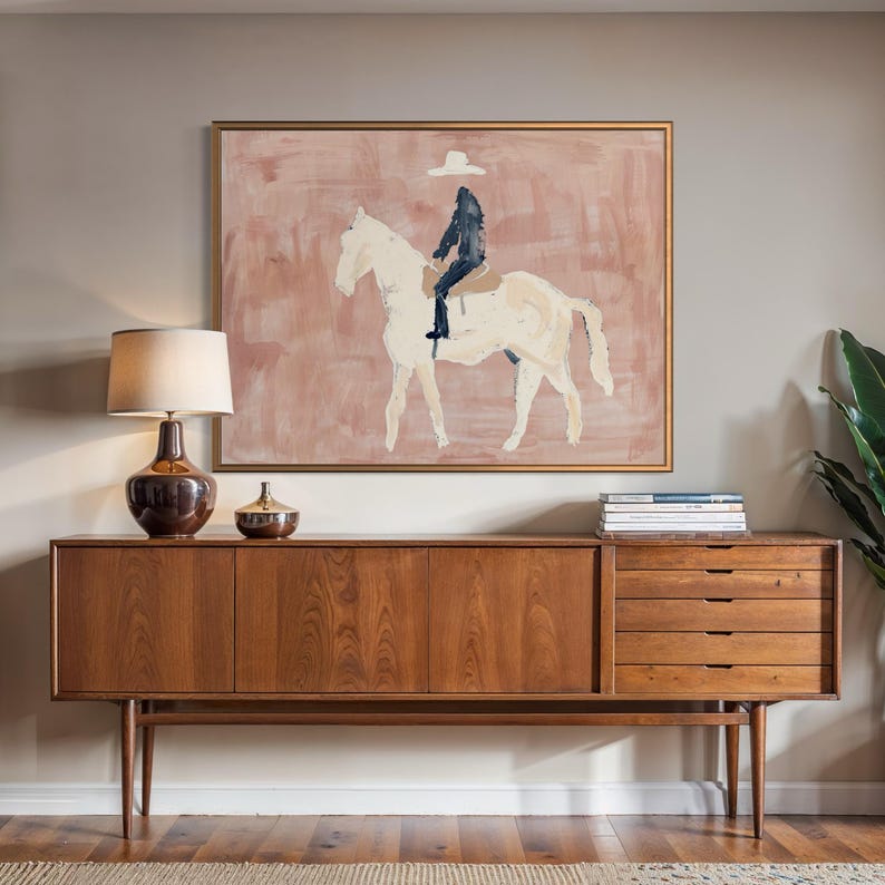 May include: A mid-century modern style wooden sideboard with a framed painting of a rider on a white horse. The painting has a pink background. A brown lamp and decorative bowl sit on the sideboard.