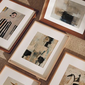 May include: A selection of framed art prints with abstract designs. The artwork includes figurative and abstract compositions. The frames are in various wood tones, and the prints are displayed on a textured surface.