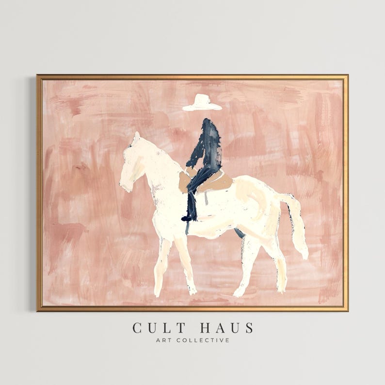 May include: Abstract art print featuring a rider on a white horse, set against a blush pink background. The rider is depicted in black attire and a white hat. The artwork is framed in a gold tone. The text "CULT HAUS ART COLLECTIVE" is at the bottom.